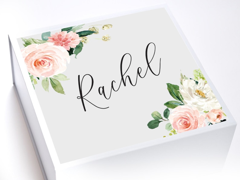 May include: A white gift box with a gray lid. The lid is decorated with a floral design in shades of pink and white. The name "Rachel" is written in black cursive script on the lid.