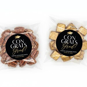 May include: Two clear cellophane bags, each containing a treat with a graduation theme. One bag holds pecans, the other gold-wrapped chocolates. Both bags feature a black circular sticker with the text "CONGRATS Grad!" and the graduate's name and class year.