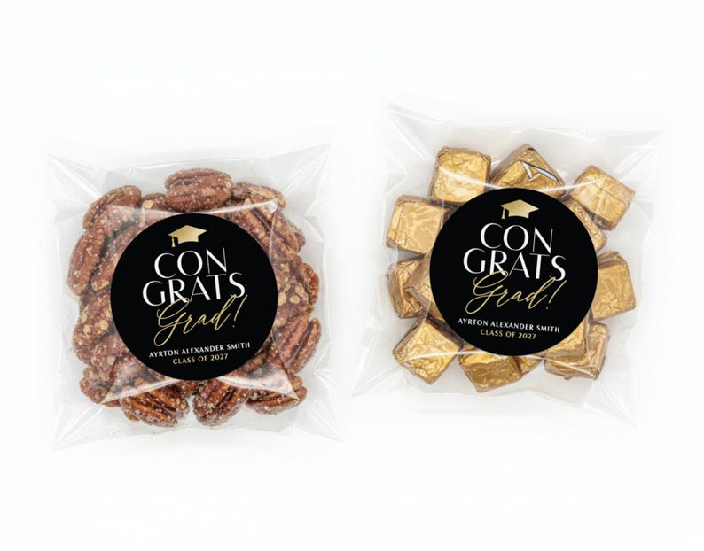 May include: Two clear cellophane bags, each containing a treat with a graduation theme. One bag holds pecans, the other gold-wrapped chocolates. Both bags feature a black circular sticker with the text "CONGRATS Grad!" and the graduate's name and class year.