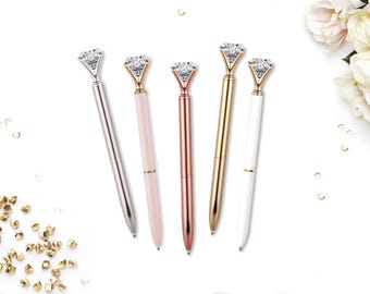 May include: Five ballpoint pens arranged in a row, each topped with a large, clear, faceted faux diamond. The pen barrels feature different finishes: silver, light pink, rose gold, gold, and white, all with metallic accents at the top.