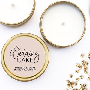 May include: Three gold candle tins with white lids. One tin has a pink label with the text "Wedding Cake" and "Smells like you're in the bridal party".