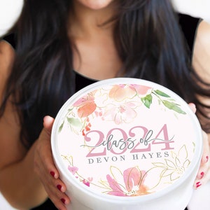 May include: White round gift box with a pink floral design and gold accents. The box has the text "Class of 2024" and "DEVON HAYES" printed on it.