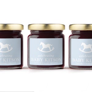 May include: Three glass jars filled with a dark red preserve, each topped with a gold lid. The jars have light blue labels with a white rocking horse and the text "celebrating BABY KITSON TAYLOR'S BABY SHOWER."