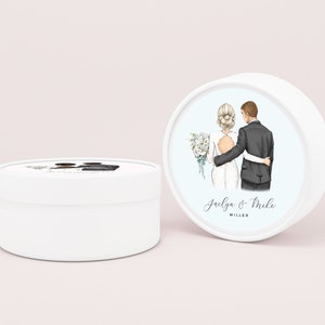 May include: White round favor box with a couple's illustration on the lid. The bride is wearing a white dress and the groom is wearing a black suit. The text on the lid reads "Jaelya & Mike MILLER".