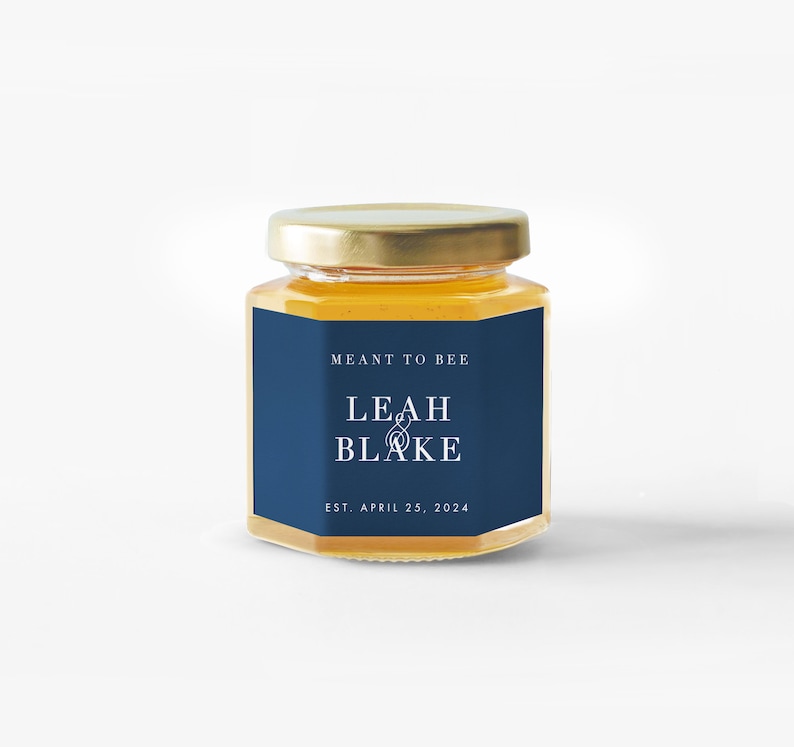 Pu&ograve; includere: Un piccolo barattolo di vetro esagonale pieno di miele, sormontato da un coperchio dorato. Un'etichetta blu navy mostra il testo "MEANT TO BEE" sopra "LEAH & BLAKE" e "EST. APRIL 25, 2024" in bianco. Il barattolo &egrave; centrato su uno sfondo bianco.