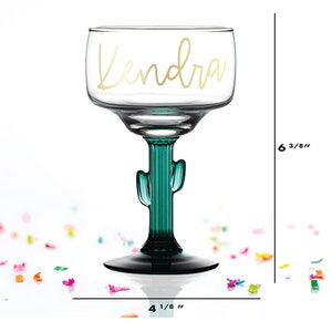 May include: A clear glass margarita glass with a green cactus stem and base. The glass is 6 3/8 inches tall and 4 1/8 inches wide. The glass has the name "Kendra" written on it in gold lettering.
