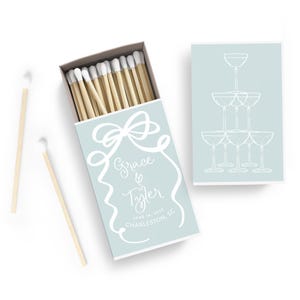 May include: A light blue matchbox with a white bow and the text "Grace & Tyler June 1st, 2023 Charleston, SC". The matchbox has a light blue background with a white design. The matchbox is open and the matches are visible. The matches are white with a wooden stick. The matchbox is sitting on a white surface. The back of the matchbox has a white design of a stack of champagne glasses.
