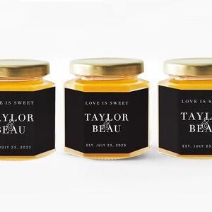 Pu&ograve; includere: Tre piccoli vasetti di vetro pieni di miele, ciascuno con coperchio dorato ed etichetta nera. L'etichetta recita "LOVE IS SWEET" sopra "TAYLOR & BEAU" e "EST. JULY 25, 2023" in testo bianco. I vasetti sono allineati.