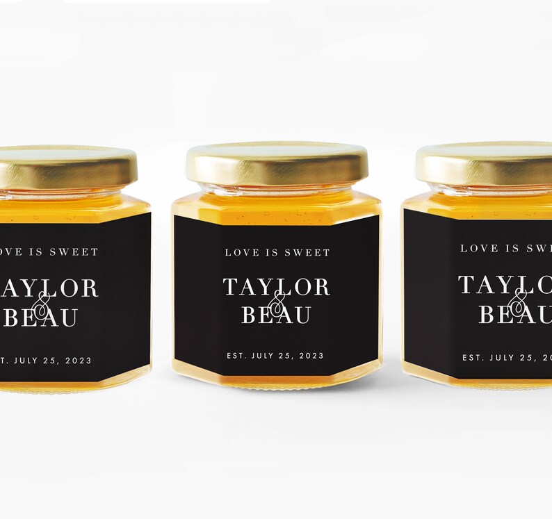 Pu&ograve; includere: Tre piccoli vasetti di vetro pieni di miele, ciascuno con coperchio dorato ed etichetta nera. L'etichetta recita "LOVE IS SWEET" sopra "TAYLOR & BEAU" e "EST. JULY 25, 2023" in testo bianco. I vasetti sono allineati.