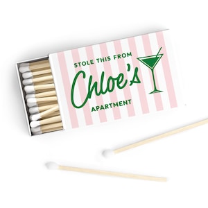 May include: A rectangular matchbox with a light pink and white vertical striped pattern. The text "STOLE THIS FROM Chloe's APARTMENT" is printed in green, accompanied by a green martini glass icon. The box contains wooden matches with white tips.