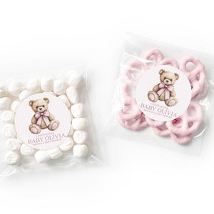 Könnte beinhalten: Zwei durchsichtige Plastiktüten mit Snacks, eine mit weißen Marshmallows und die andere mit rosa schokolierten Brezeln. Jede Tüte hat einen runden Aufkleber mit einer braunen Teddybär-Illustration, einer rosa Schleife und dem Text "celebrating BABY OLIVIA MONICA'S BABY SHOWER".