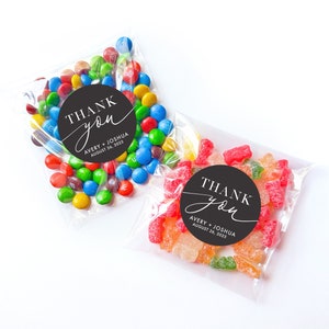 Peut inclure: Deux sachets en plastique transparent remplis de bonbons. Un sachet contient des M&Ms color&eacute;s et un autocollant rond noir et blanc qui dit "THANK you" avec le texte "AVERY + JOSHUA AUGUST 26, 2025" en dessous. L'autre sachet contient des bonbons g&eacute;lifi&eacute;s color&eacute;s et un autocollant rond noir et blanc qui dit "THANK you" avec le texte "AVERY + JOSHUA AUGUST 26, 2025" en dessous.