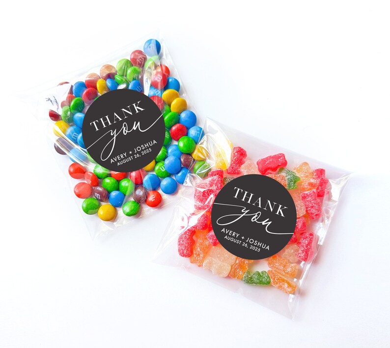 Peut inclure: Deux sachets en plastique transparent remplis de bonbons. Un sachet contient des M&Ms color&eacute;s et un autocollant rond noir et blanc qui dit "THANK you" avec le texte "AVERY + JOSHUA AUGUST 26, 2025" en dessous. L'autre sachet contient des bonbons g&eacute;lifi&eacute;s color&eacute;s et un autocollant rond noir et blanc qui dit "THANK you" avec le texte "AVERY + JOSHUA AUGUST 26, 2025" en dessous.