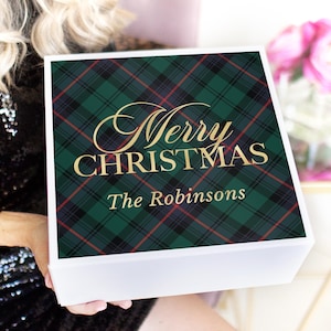 Könnte beinhalten: Eine quadratische weiße Geschenkbox mit einem grün-schwarz-roten Schottenkaro-Muster auf dem Deckel. Der Deckel zeigt den goldenen Schriftzug Merry CHRISTMAS und The Robinsons in einer Kombination aus Schreib- und Serifenschrift.