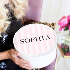 May include: A white round gift box with a pink and white striped lid. The lid has the name "SOPHIA" printed in black.
