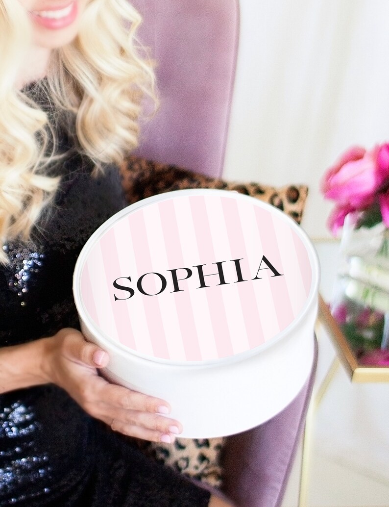 May include: A white round gift box with a pink and white striped lid. The lid has the name "SOPHIA" printed in black.