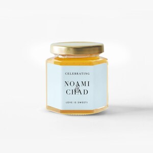 Pu&ograve; includere: Un piccolo barattolo di vetro esagonale pieno di miele dorato, sormontato da un coperchio dorato. Un'etichetta azzurra mostra i nomi "NOAMI & CHAD" con il testo "CELEBRATING" sopra e "LOVE IS SWEET!" sotto.