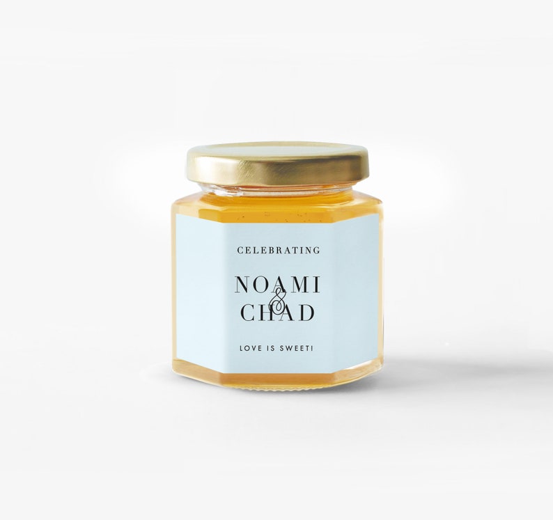 Pu&ograve; includere: Un piccolo barattolo di vetro esagonale pieno di miele dorato, sormontato da un coperchio dorato. Un'etichetta azzurra mostra i nomi "NOAMI & CHAD" con il testo "CELEBRATING" sopra e "LOVE IS SWEET!" sotto.