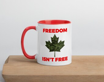 May include: White ceramic mug with a red rim and handle. The mug features a green camouflage maple leaf and the text "FREEDOM ISN'T FREE" in red.