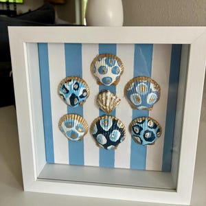 May include: A square white shadow box frame containing seven seashells arranged on a light blue and white striped background. Each shell is individually painted with blue, white, and gold patterns, featuring various spots and circular designs.
