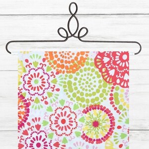 May include: A colorful floral patterned fabric displayed on a decorative black metal hanger. The fabric features a repeating design of flowers and dots in shades of red, pink, orange, yellow, green, and blue against a white background.