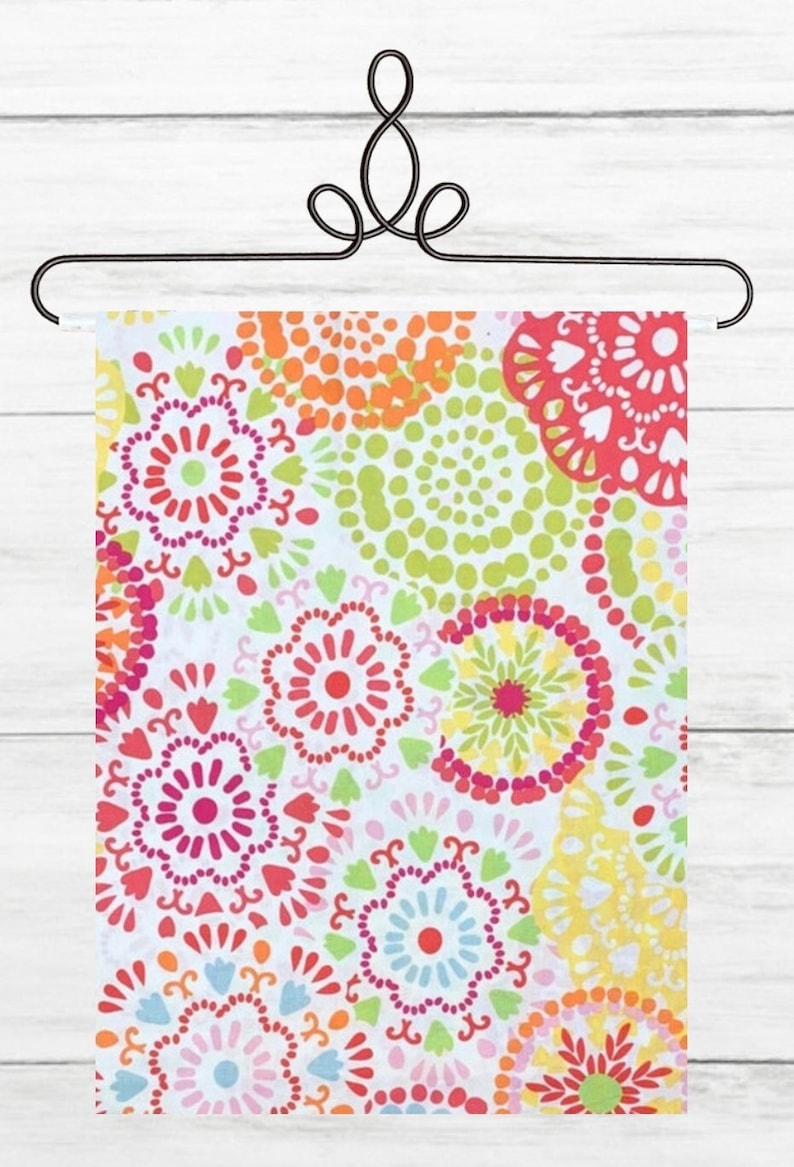 May include: A colorful floral patterned fabric displayed on a decorative black metal hanger. The fabric features a repeating design of flowers and dots in shades of red, pink, orange, yellow, green, and blue against a white background.