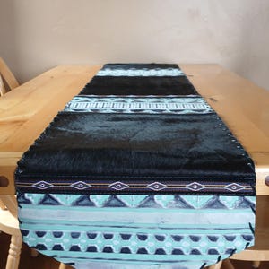 May include: A rectangular table runner with a black faux fur center and patterned ends. The ends feature a geometric design in shades of teal, white, and black, with a stitched border. The runner is placed on a wooden table.
