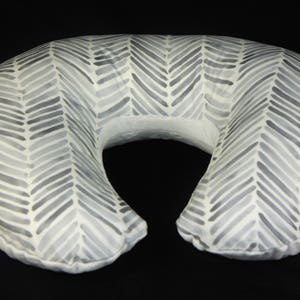May include: A gray and white chevron patterned nursing pillow. The pillow is shaped like a crescent moon and has a hole in the middle for the user's head.