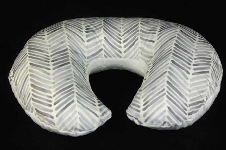 May include: A gray and white chevron patterned nursing pillow. The pillow is shaped like a crescent moon and has a hole in the middle for the user's head.