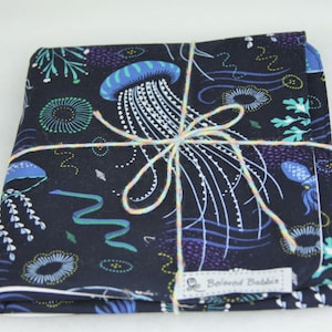 May include: A black fabric baby burp cloth with a colorful jellyfish and seaweed pattern. The cloth is tied with a light pink and yellow string and has the text "Beloved Babble" on the bottom right corner.