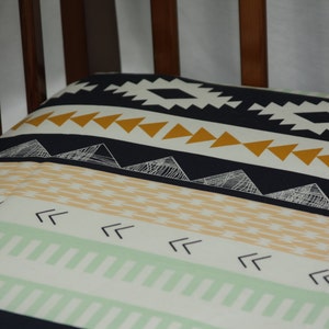 May include: A crib sheet with a geometric pattern in shades of green, yellow, white, and black. The pattern features triangles, arrows, and lines.