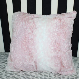May include: A soft pink and white faux fur pillow with a fluffy texture. The pillow is on a gray and white patterned surface.
