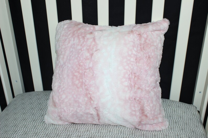 May include: A soft pink and white faux fur pillow with a fluffy texture. The pillow is on a gray and white patterned surface.