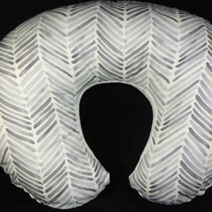 May include: A white and gray chevron patterned nursing pillow. The pillow is shaped like a crescent moon and has a hole in the center for the baby's head.
