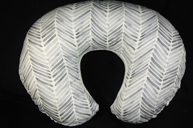 May include: A white and gray chevron patterned nursing pillow. The pillow is shaped like a crescent moon and has a hole in the center for the baby's head.
