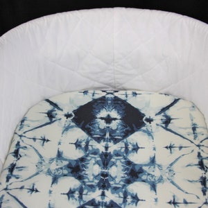 May include: A white quilted bassinet with a blue and white tie-dye patterned fitted sheet.