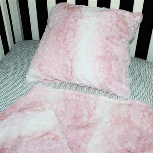 May include: A pink and white faux fur crib blanket and pillow set. The blanket and pillow are both soft and fluffy.