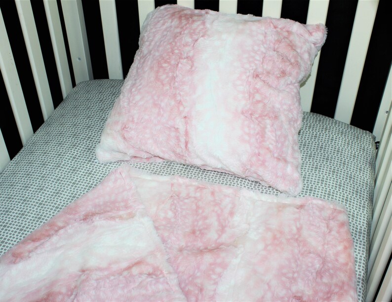 May include: A pink and white faux fur crib blanket and pillow set. The blanket and pillow are both soft and fluffy.