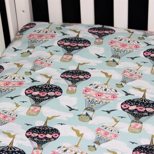Peut inclure: Un drap de lit pour bébé de couleur bleu clair avec un motif de montgolfières aux motifs floraux. Les montgolfières sont noires, blanches et roses avec des accents dorés. Le fond est bleu clair avec des nuages blancs.