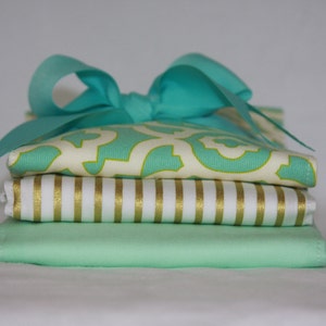 May include: A stack of four fabric squares in shades of green and white. The top square has a teal and white geometric pattern. The second square has white and gold stripes. The third square is solid light green. The bottom square is a darker shade of green. A teal ribbon is tied around the stack.