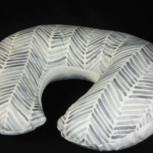 May include: A white and gray patterned nursing pillow shaped like a crescent moon. The pillow has a chevron pattern with gray lines on a white background.