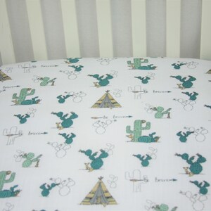 May include: White fitted crib sheet with a teal cactus and teepee pattern. The sheet has a repeating design of cacti, teepees, and the phrase "be brave" in a handwritten font. The sheet is made of a soft, breathable fabric.