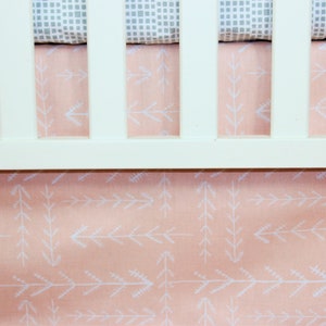 May include: A white crib with a coral-colored crib skirt featuring a white arrow pattern. The crib sheet is gray with a white square pattern. The crib is a nursery item.