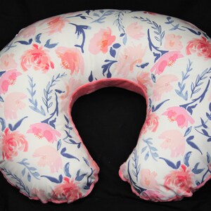 May include: A white and pink floral print nursing pillow with a pink trim. The pillow is shaped like a crescent moon and is designed to support a pregnant woman or a new mother while breastfeeding.