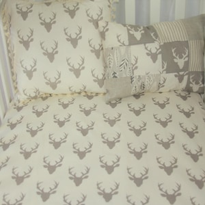 May include: A white crib with a beige fitted sheet featuring a repeating pattern of grey deer silhouettes. Two decorative pillows are on the crib, one with a patchwork design and the other with a repeating pattern of grey deer silhouettes.