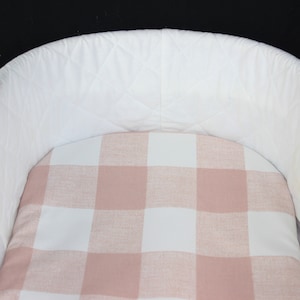 Puede incluir: Una cuna blanca con exterior acolchado e interior con estampado de cuadros rosas y blancos. La cuna tiene forma curva y está diseñada para bebés. Los colores suaves y neutros la hacen adecuada para cualquier guardería.