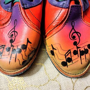 Puede incluir: Un par de zapatos de cuero rojo con un degradado arco&iacute;ris y notas musicales negras pintadas en los dedos. Los zapatos tienen detalles morados alrededor del tobillo.