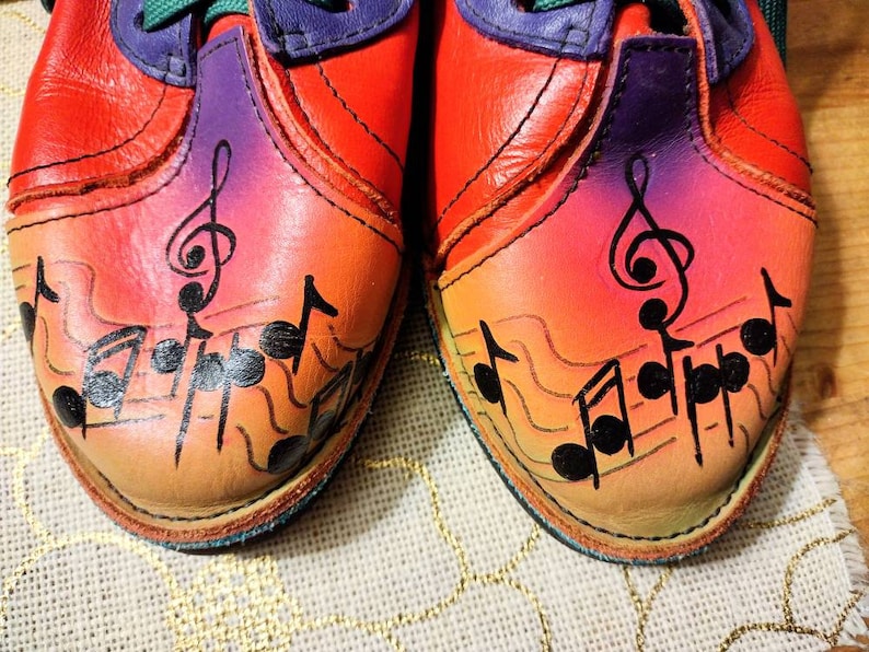 Puede incluir: Un par de zapatos de cuero rojo con un degradado arco&iacute;ris y notas musicales negras pintadas en los dedos. Los zapatos tienen detalles morados alrededor del tobillo.