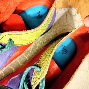 Puede incluir: Un par de zapatos de cuero coloridos con cordones verdes. Los zapatos tienen un dise&ntilde;o morado, amarillo y rojo con detalles azules. El interior de los zapatos tiene una plantilla azul con la palabra "Nuf" escrita en ella.