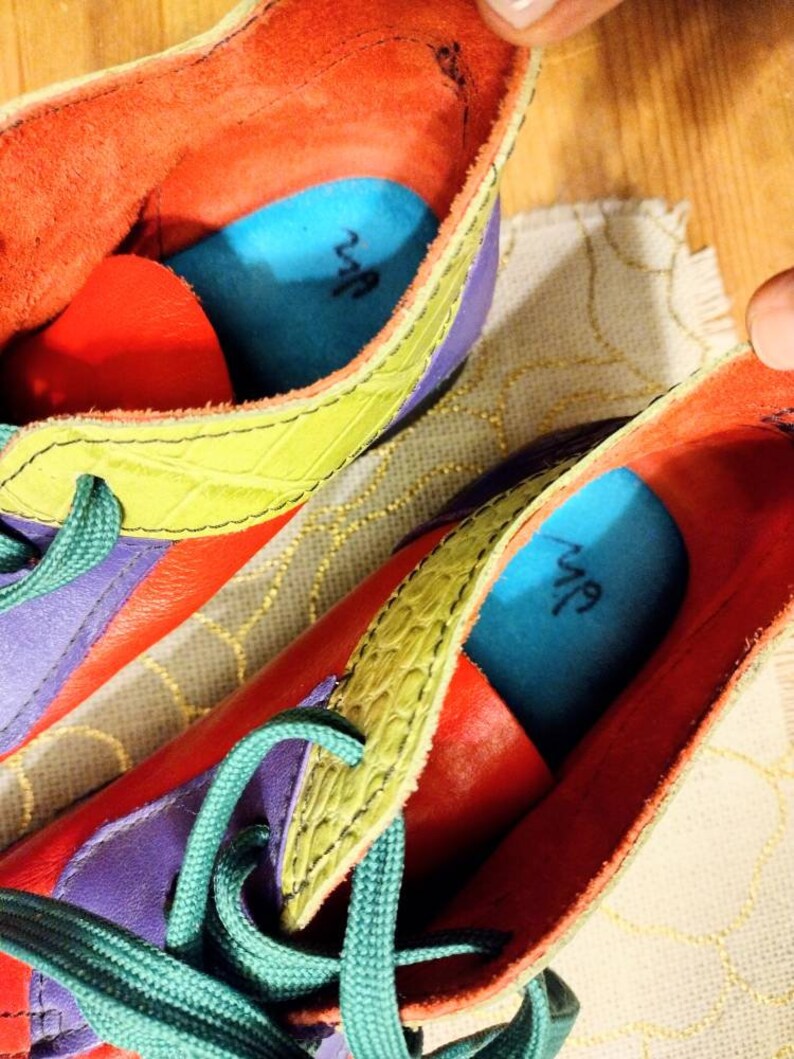 Puede incluir: Un par de zapatos de cuero coloridos con cordones verdes. Los zapatos tienen un dise&ntilde;o morado, amarillo y rojo con detalles azules. El interior de los zapatos tiene una plantilla azul con la palabra "Nuf" escrita en ella.